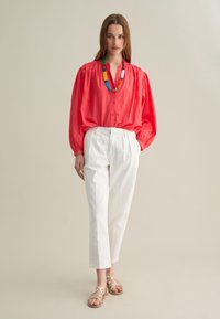 Blusa rossa a maniche lunghe con bottoni, pantaloni bianchi a vita alta e sandali beige con cinturini. Presenta una collana colorata di perline.