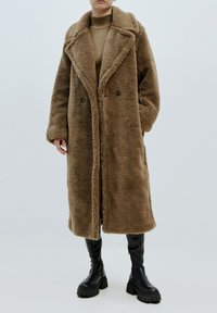 Brauner, flauschiger Kunstfellmantel mit breitem Kragen, doppelter Knopfleiste und Seitentaschen. Getragen über einem gerippten beigen Pullover und kombiniert mit schwarzen Stiefeln.