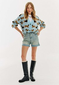 Mladá žena v modrém kardiganu s vzorem psů, džínových šortkách a černých kozačkách nad kolena stojí se založenýma rukama v bok.