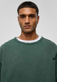 Groene sweatshirt met een iets versleten halslijn en een klein gaatje in de mouw. Gemaakt van zachte stof, met een comfortabele pasvorm en een gestructureerd oppervlak.