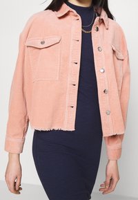 Różowa kurtka ze sztruksu z dwoma przednimi kieszeniami, srebrnymi guzikami i krótkim krojem. Materiał o fakturze z lekko postrzępionym dołem.