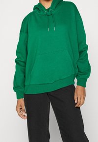 Sweat à capuche vert en tissu doux, doté d'une poche avant, de manches longues, d'une coupe décontractée et de poignets et ourlet côtelés.