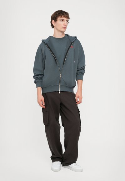 Un hoodie zippé gris superposé sur un T-shirt noir, associé à un pantalon cargo noir ample. Des baskets blanches complètent la tenue, mettant l'accent sur le confort.