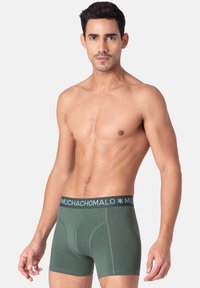 Zelena bombažna bokserica s temno modrim pasom, na katerem je napis blagovne znamke "MUCHACHOMALO" v svetlo modri barvi. Gladka tekstura, prilegajoča zasnova.