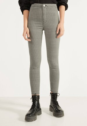 Person wearing black and white checkered slim-fit pants and black lace-up chunky boots standing on a white floor.