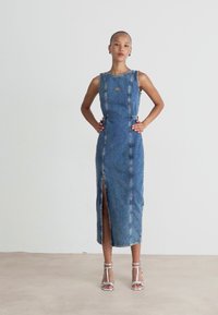 Denim midi haljina s bočnim izrezom, podesivim naramenicama, izblijedjelim plavim opranjem i kontrastnim šavovima. Model nosi srebrne sandale na remen.