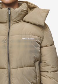 Puhasti kaput na svijetlom bež materijalu s visokim ovratnikom, zatvaranjem na zip i istaknutim "PEGADOR" logotipom. Sadrži quilted dijelove za izolaciju.