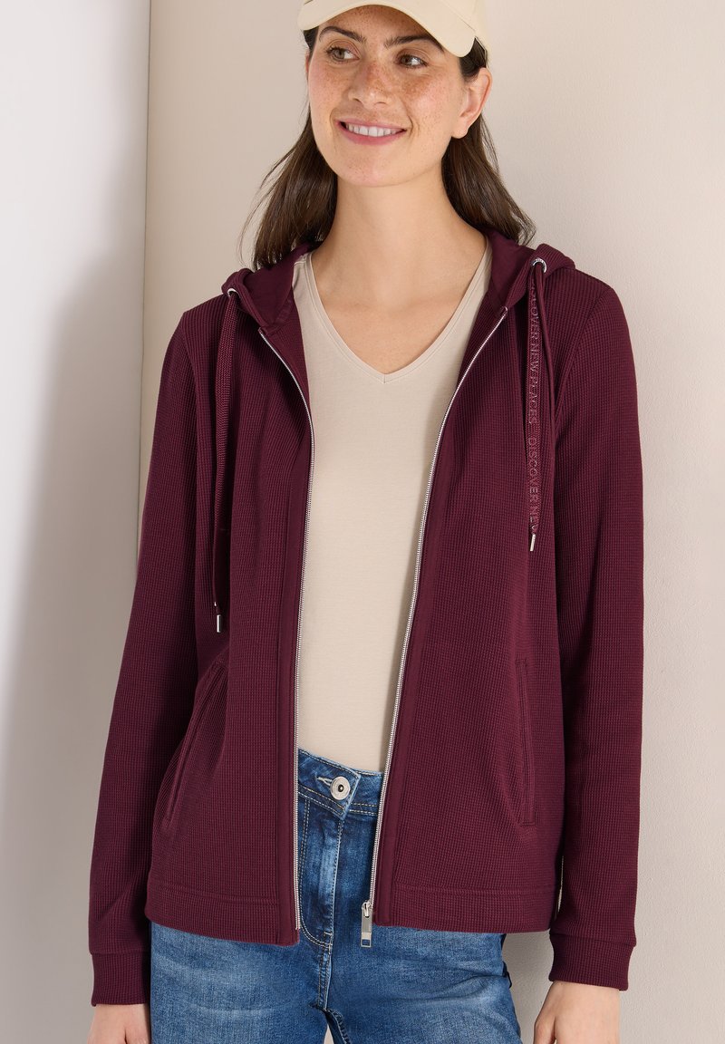 Mujer sonriente con gorra beige, camisa beige de cuello en V, sudadera granate sin cremallera y jeans azules, de pie frente a una pared lisa.