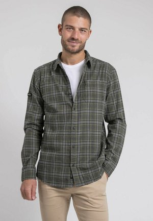 Chemise en flanelle à carreaux verts, à manches longues, avec un devant boutonné et une poche poitrine. Portée par-dessus un T-shirt blanc.