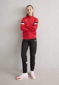 Rdeča jakna z zadrgo in črno-belimi črtami, združena s črnimi športnimi hlačami in belimi čevlji z oranžnimi poudarki.