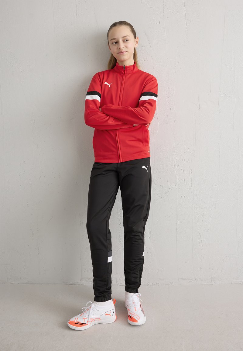 Rdeča jakna z zadrgo in črno-belimi črtami, združena s črnimi športnimi hlačami in belimi čevlji z oranžnimi poudarki.