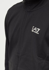 Giacca nera con zip realizzata in tessuto morbido e elasticizzato. Presenta un colletto alto e il logo EA7 sul lato sinistro del petto. Texture liscia con un design aderente.
