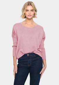 Hellrosa Strickpullover mit weitem Ausschnitt, Dreiviertelärmel und gerippter Textur, kombiniert mit dunkelblauen Jeans mit Fronttaschen.