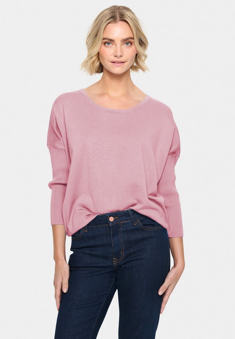 Hellrosa Strickpullover mit weitem Ausschnitt, Dreiviertelärmel und gerippter Textur, kombiniert mit dunkelblauen Jeans mit Fronttaschen.