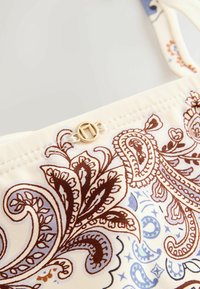 Gros plan sur un tissu avec des motifs cachemire marron et bleu et un petit fermoir rond doré sur une bretelle de vêtement couleur crème.