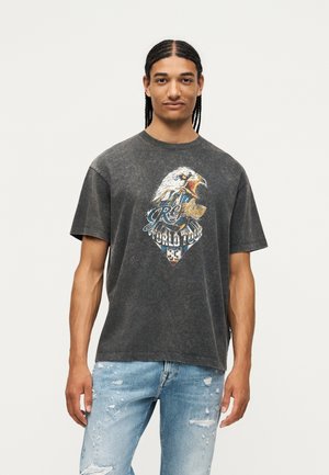 Joven con cabello trenzado que lleva una camiseta negra desgastada con un gráfico de águila, "Replay World Tour", y vaqueros azul claro rasgados.