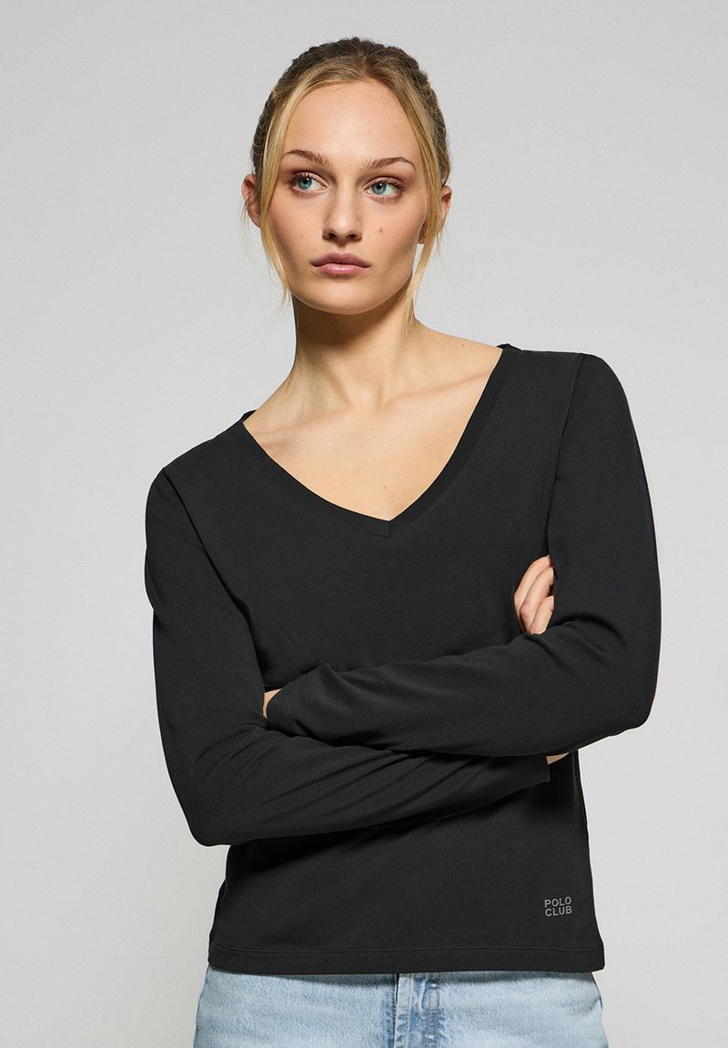 Svart långärmad V-ringad topp i ribbat tyg, med en subtil logga i nedersta hörnet, ihop med ljusblå jeans.