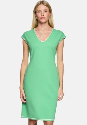 Woman standing facing forward wearing a sleeveless green knee-length dress with white trim along neckline and hem.