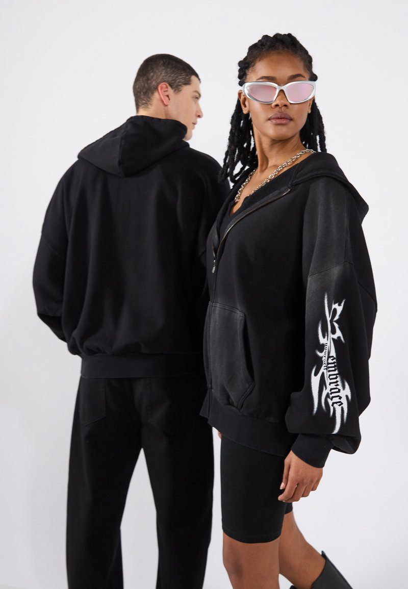 Black zip-up hoodie featuring front pockets and a flame graphic on the right sleeve; paired with black bike shorts. Lightweight fabric, relaxed fit.