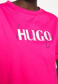 Osoba nosząca jasnoróżową koszulkę z krótkim rękawem, z wyraźnym białym i czarnym napisem "HUGO" na środku klatki piersiowej.