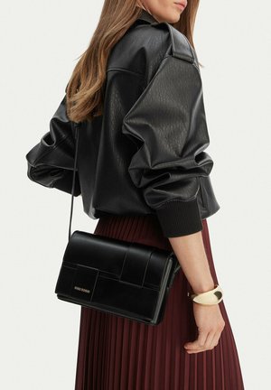 Frau trägt schwarze Lederjacke und plissierten dunkelroten Rock, trägt schwarze Schultertasche und goldenes Armspangenarmband am Handgelenk.