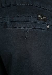 Jeans de mezclilla azul oscuro que presentan un bolsillo trasero con detalles de costura y un parche con el logo. Incluyen un botón de metal y un patrón de costura distintivo.