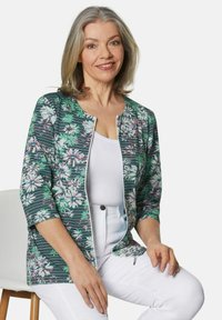 Blumenmuster-Jacke in Grün, Weiß und Rosa mit einem Reißverschluss vorne. Verfügt über 3/4-Ärmel und einen leichten, strukturierten Stoff. Wird über einem weißen Oberteil getragen.