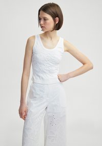 Weißes ärmelloses Oberteil mit strukturiertem besticktem Muster, kombiniert mit weißen weit geschnittenen Hosen mit floralen Ausschnitten.