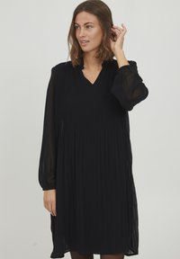 Schwarzes, plissiertes Kleid mit V-Ausschnitt, langen transparenten Ärmeln und Rüschendetail am Ausschnitt. Der Stoff wirkt leicht und fließend.