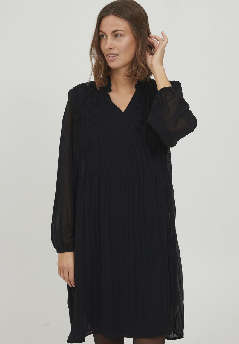 Schwarzes, plissiertes Kleid mit V-Ausschnitt, langen transparenten Ärmeln und Rüschendetail am Ausschnitt. Der Stoff wirkt leicht und fließend.