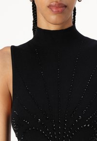 Top fără mâneci, cu guler înalt, de culoare neagră, prezentând un model texturat de paiete negre aranjate într-un design în evantai, realizat dintr-un material tricotat moale.