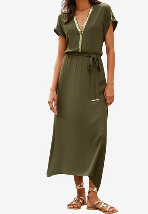 Femme portant une robe maxi vert olive avec un bord doré au niveau du col en V et des épaules, ceinture nouée à la taille, et sandales plates dorées à brides.