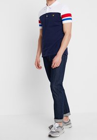Navy and white polo shirt with red and blue striped sleeves, yellow logo, paired with dark denim jeans and gray sneakers. Casual fit.