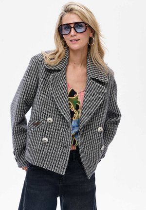 Frau mit übergroßen schildpattfarbenen Sonnenbrillen, Ohrreifen, grauem Hahnentrittmuster-Doppelreiherjacke, buntem gemustertem Oberteil und dunkler Hose.