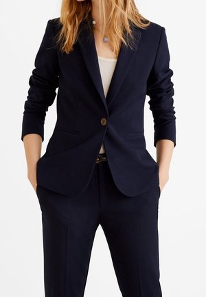 Femme portant un blazer ajusté bleu marine et un pantalon assorti avec un haut blanc en dessous, les mains dans les poches, sur un fond blanc.