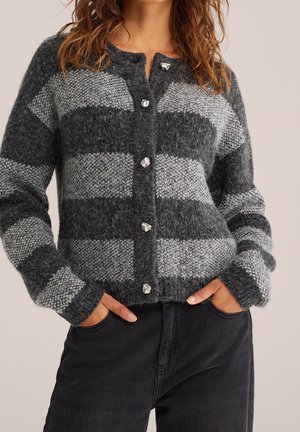 Femme portant un cardigan gris rayé duveteux avec des boutons argentés et un pantalon noir, debout les mains dans les poches.