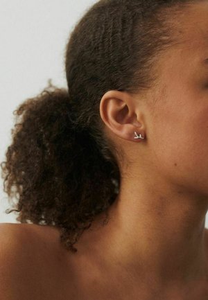 Tuva personāža ar cirtainiem matiem, kas sasieti atpakaļ, kreisajā ausī nēsā nelielu sudraba auskaru, kas veidots kā zariņš.