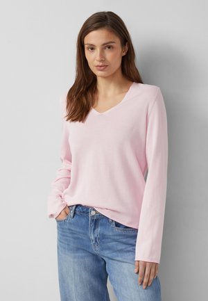 Frau mit braunen Haaren, die ein hellrosa langärmliges V-Ausschnitt-Shirt und blaue Jeans trägt, steht mit einer Hand in der Tasche vor einer grauen Wand.