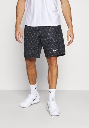 Mężczyzna noszący czarne spodenki Nike z wzorem diamentu, białe skarpetki Nike, białe buty sportowe Nike i białą sportową koszulkę.