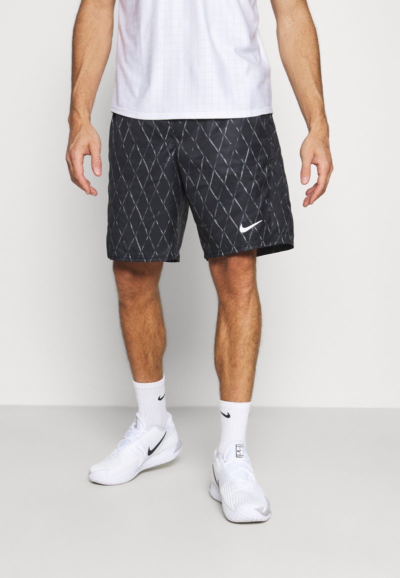 Homme portant un short noir Nike avec un motif en losange, des chaussettes blanches Nike, des baskets blanches Nike et un t-shirt de sport blanc.