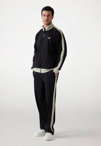 Survêtement de sport noir avec des rayures latérales crème. Veste zippée avec col montant, pantalon assorti et baskets blanches. Texture de tissu lisse.