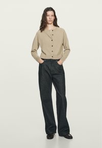 Cardigan in misto lana beige con scollatura rotonda e bottoni neri, abbinato a jeans a gamba larga in denim scuro. Tessuto morbido, vestibilità casual.