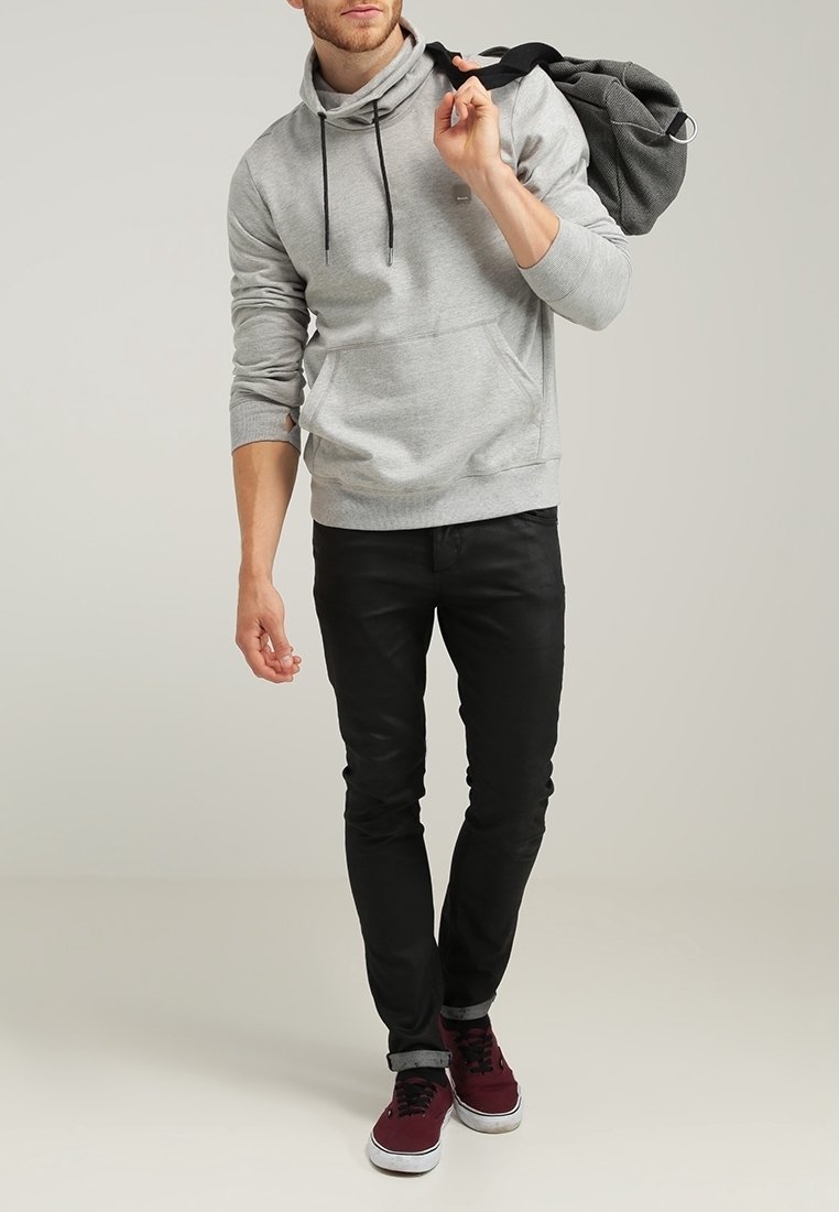 Homme portant un sweat à capuche gris, un pantalon noir et des baskets bordeaux, portant une veste grise sur l'épaule en marchant vers l'avant.