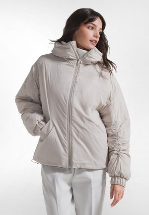 Femme portant une doudoune zippée beige clair avec des manches froncées et un col montant, associée à un pantalon blanc large, sur fond blanc.