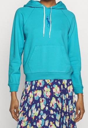 Personne portant un sweat à capuche turquoise avec des cordons blancs et une jupe plissée à motif floral dans des tons de bleu, vert, blanc et rose.