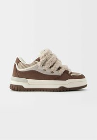 Sneaker i en beige och brun färgsättning. Har en nätövre, mjuka tygremmar och en gummisula med texturerad detalj.