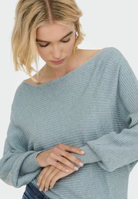Šviesiai mėlynas raštuotas ilgos rankovės megztinis su nuogais pečiais ir tekstūruotu audiniu. Modelio rankos švelniai laiko megztinio apvadą.