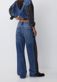 Persona vistiendo una chaqueta de mezclilla a dos tonos, azul y negro, y unos jeans de pierna ancha a juego, de pie contra un fondo blanco liso.