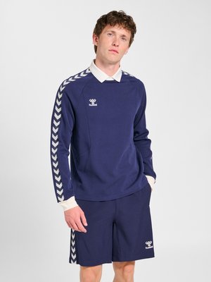 Joven viste ropa deportiva azul marino con rayas blancas en forma de cheurón en las mangas y los pantalones cortos, de pie frente a un fondo liso de color gris claro.