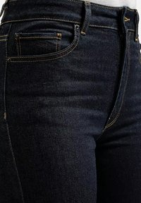 Granatowe dżinsy z widocznymi kieszeniami z przodu i na monety, pomarańczowymi kontrastowymi szwami i pętlami na pasek noszone w talii.